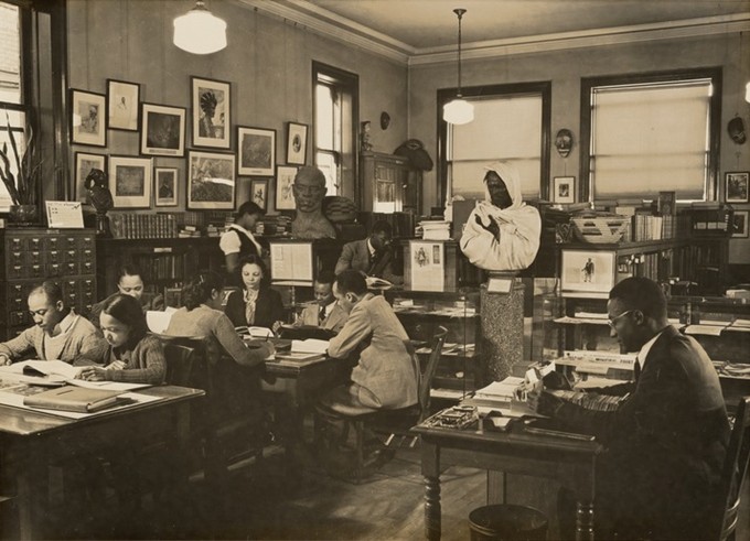 A picture of the 135th street branch of the New York Public Library. Schomburg Center reading room. It is black and white, showing a small room crowded with several tables and other furniture.  A number of people appearing to be of African American and Asian descent are sitting looking at the collection materials.